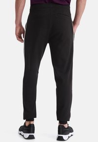 Schwarze Jogginghose mit taillierter Form, seitlichen Streifen, elastischem Bund und einer Gesäßtasche. Kombiniert mit schwarzen Sportschuhen.