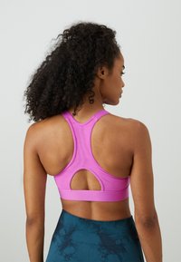 Frau mit lockigem Haar, die einen pinken Sport-BH mit Racerback und dunkelblauen gemusterten Leggings trägt, mit dem Rücken zu einer schlichten Wand.