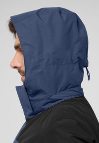 Veste à capuche bleu foncé avec une partie inférieure texturée noire. Dotée d'un col haut et d'un cordon de serrage pour ajuster la capuche.