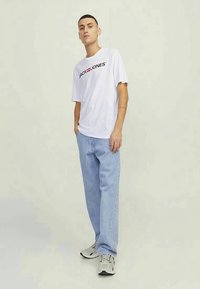 Νεαρός άνδρας όρθιος, φορώντας λευκό T-shirt "Jack & Jones", ανοιχτό μπλε τζιν και γκρι αθλητικά παπούτσια, σε ένα φωτεινό ουδέτερο φόντο.