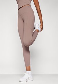 Light brown leggings with a high waistband, featuring side pockets and stitched detailing, paired with white athletic shoes.