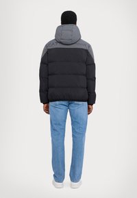 Giacca a vento nera e grigia con cappuccio, design a righe orizzontali e dettaglio del logo sul retro. Indossata sopra jeans blu e scarpe bianche.