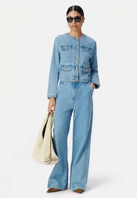 Jasnoniebieska kurtka dżinsowa z złotymi guzikami i czterema przednimi kieszeniami, zestawiona z szerokimi jasnoniebieskimi dżinsowymi spodniami. Modelka trzyma beżową torbę.