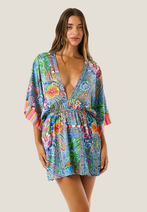 Giovane donna che indossa un abito colorato con motivo floreale e paisley, con una profonda scollatura a V e maniche larghe, in piedi davanti a uno sfondo semplice.