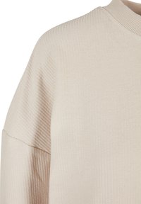 Pull beige côtelé avec un col montant et une coupe ample. Le tissu présente une apparence douce et texturée avec des épaules tombantes décontractées.