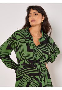 Grünes und schwarzes gemustertes Hemdkleid aus glattem Stoff, mit langen Ärmeln, Knopfschluss und einem Bindegürtel in der Taille.