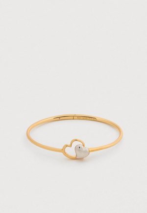 Bracelet jonc en métal doré avec un design de cœur entrelacé et un accent en forme de cœur blanc. Texture lisse, style ouvert.