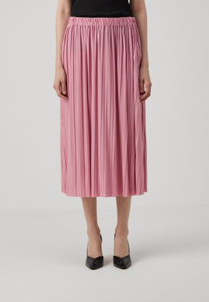 Kvinde iført en pink plisseret midi-nederdel med elastisk taljebånd og sorte spidse hæle, stående mod en ensfarvet baggrund.