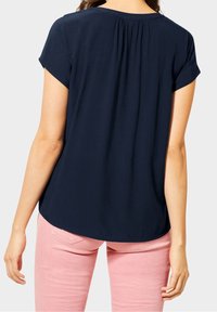 Marineblauwe blouse met korte mouwen en een ronde halslijn, met plooidetail op de rug; gedragen met lichtroze broek.