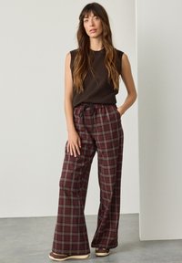 Weit geschnittene, bordeauxrote, karierte Hose mit elastischem Bund und Kordelzug. Kombiniert mit einem ärmellosen, dunkelbraunen Oberteil. Legerer Stoff mit Struktur.