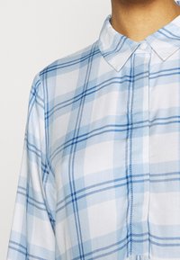 Camicia a quadri blu e bianca con colletto. Tessuto di cotone, caratterizzata da strisce sottili e spesse in un motivo a scacchi. Chiusura a bottoni al collo.
