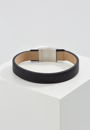 Bracelet en cuir noir avec doublure intérieure beige et fermoir métallique argenté rectangulaire, présenté sur un socle blanc sur fond gris.