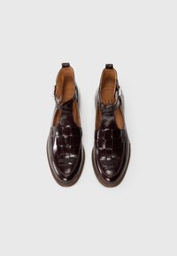 Brown leather shoes with a woven upper design, open heel, and ankle strap. Glossy finish with round toe and contrasting sole stitching.