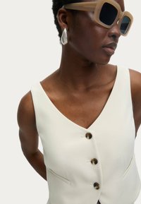 Ivory mouwloze vest met een V-hals, voorzien van drie ronde, donkere knopen en twee zijzakken, met een gladde stof en een minimalistisch ontwerp.