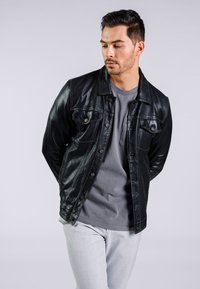 Chaqueta de cuero negra con bolsillos frontales y cierres de botón, combinada con una camiseta gris. Modelo vistiendo pantalones gris claro, fondo neutro.
