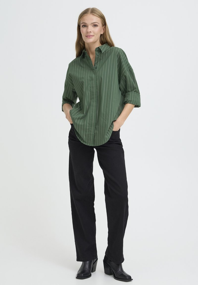Grünes gestreiftes Hemd mit Button-Down-Kragen, hochgerollten Ärmeln und lockerer Passform, kombiniert mit schwarzen geraden Hosen und schwarzen Stiefeln.