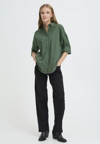 Camicia a righe verdi con colletto button-down, maniche arrotolate e vestibilità ampia, abbinata a pantaloni neri a gamba dritta e stivali neri.