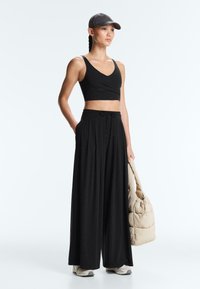 Žena v černém krátkém topu, širokých kalhotách, šedé čepici a bílých teniskách, držící béžovou polstrovanou tašku, stojící na bílém pozadí.