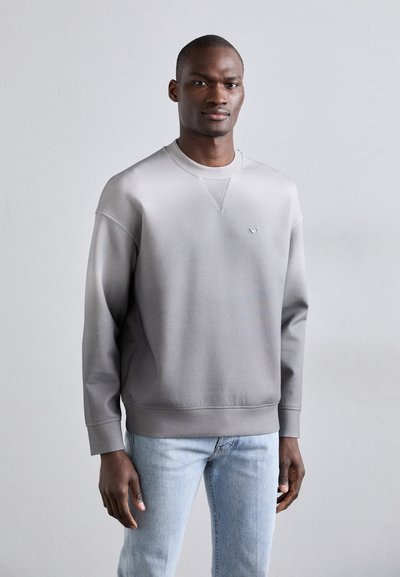 Gray sweatshirt with a textured finish, crew neck, and subtle logo on the chest, paired with light blue jeans. Smooth fabric and relaxed fit.