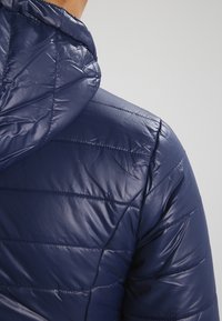 Granatowa kurtka puchowa z kapturem, błyszczącą teksturą, pionowymi przeszyciami i elastycznymi mankietami. Materiał wydaje się lekki i izolowany.