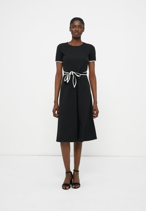 Γυναίκα που φοράει μαύρο φόρεμα μήκους μέχρι το γόνατο με λευκή λεπτομέρεια στα μανίκια και δεμένη λευκή ζώνη, συνδυασμένο με μαύρα πέδιλα με λουράκι στον αστράγαλο, στέκεται μπροστά σε λευκό τοίχο.