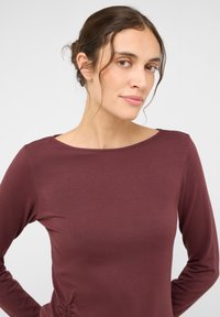 Langarm-Top in Burgund, mit rundem Ausschnitt und strukturiertem Stoff sowie dezentem Raffdetail an der Seite für eine taillierte Silhouette.