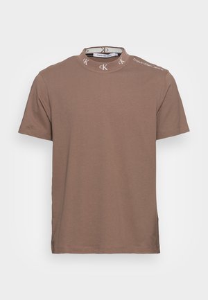 Brązowy t-shirt z krótkim rękawem i kołnierzem typu mock neck z powtarzającym się białym napisem "cK" i "Calvin Klein Jeans" na ramieniu.