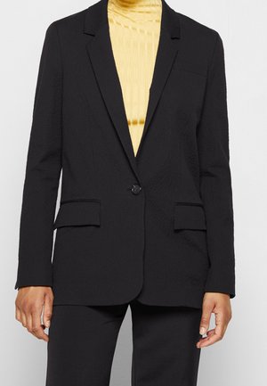 Zwarte blazer met één knoop en ingesneden revers, gedragen over een geribbelde gele coltrui en bijpassende zwarte broek.