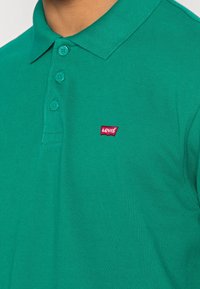 Zielona koszulka polo wykonana z teksturowanej tkaniny, z kołnierzykiem i trzema guzikami. Zauważalna czerwona naszywka z logo Levi's na lewej części klatki piersiowej.