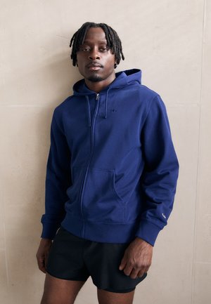 Haut de hoodie zippé bleu marine en tissu mélange coton, avec une poche avant, un capuchon avec cordon de serrage et un détail de logo sur la poitrine.