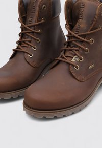 Bottes montantes en cuir marron avec des semelles robustes, œillets en métal et étiquettes de marque sur la languette et sur le côté, conçues pour être portées à l'extérieur.