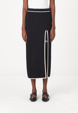 Femme portant une jupe midi noire avec un bord blanc et une fente verticale à l'avant droite, associée à des chaussures noires à boucle et un haut blanc.