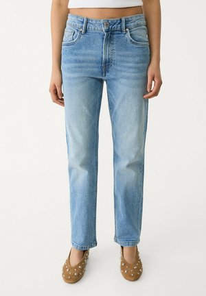 Vrouw draagt lichtblauwe rechte jeans en bruine platte schoenen met zilveren studs, staand tegen een effen witte achtergrond.
