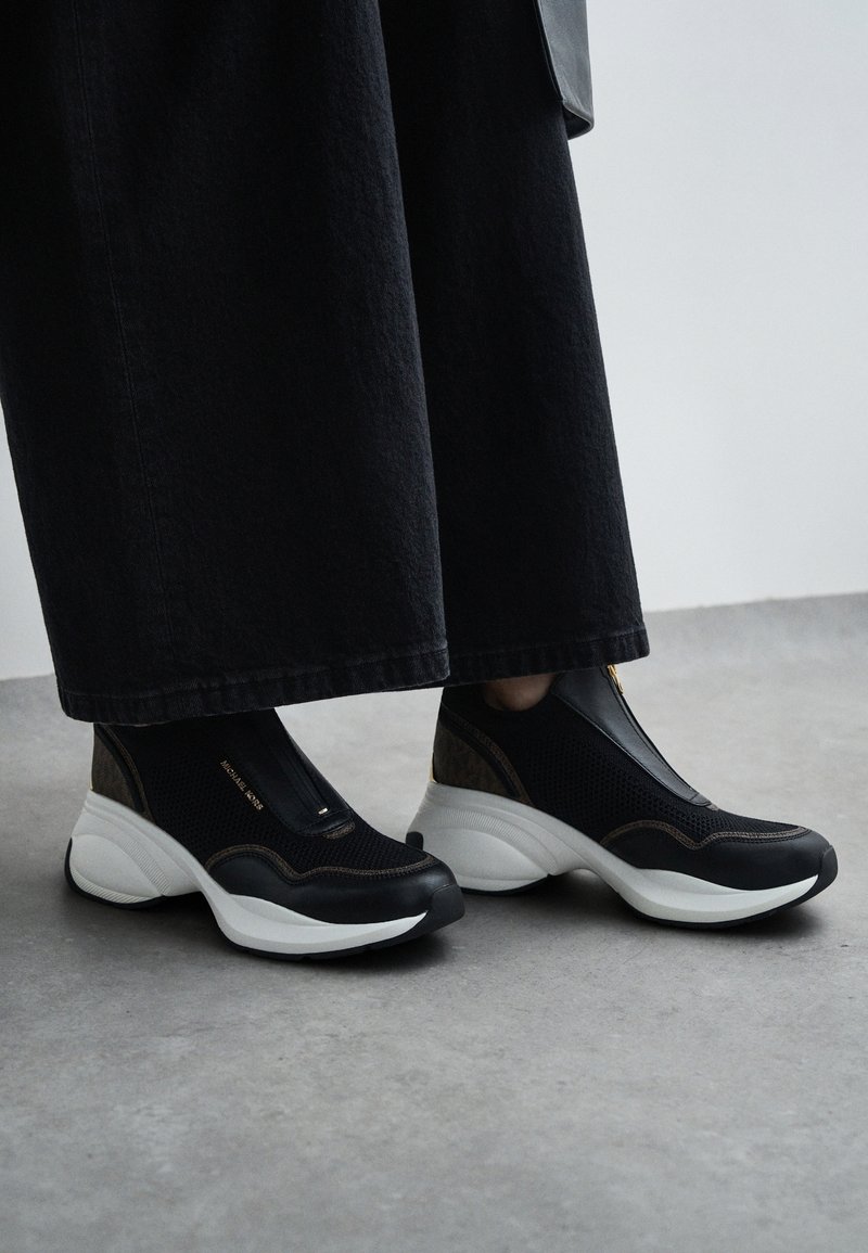 Baskets slip-on noir et blanc à semelles épaisses portées avec un pantalon noir raccourci sur un sol gris.