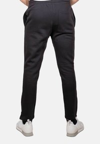 Uomo che indossa pantaloni da jogging neri e sneakers bianche, mostrato di schiena su uno sfondo semplice.