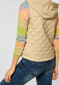 béžová prošívaná vesta s kapucí, která má barevné kostkované vzory na rukávech v pastelových odstínech. Každý rukáv má žebrovaný manžet.