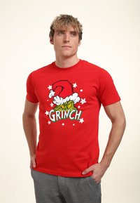 Mężczyzna w czerwonej koszulce z nadrukiem kreskówkowej twarzy Grincha w czapce Mikołaja i białymi gwiazdkami, stoi z rękami w kieszeniach, patrząc lekko w lewo.
