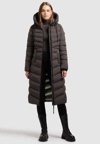 Lang grå pufferfrakke med hætte, der har quiltede paneler, en lynlås fortil og en lysere kontrastforing. Styliseret med sorte leggings og støvler.