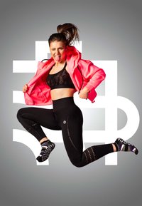 Modelka w aktywnej odzieży sportowej w jasnoróżowej kurtce, czarnym, teksturowanym biustonoszu sportowym, czarnych legginsach z wysokim stanem i wzorzystych skarpetkach, skacze na szarym tle.