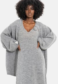 Szary, oversize'owy, dziergany kardigan z dekoltem w serek, noszony na szarą sukienkę. Kardigan ma teksturowane rękawy i ozdobne guziki na mankietach.