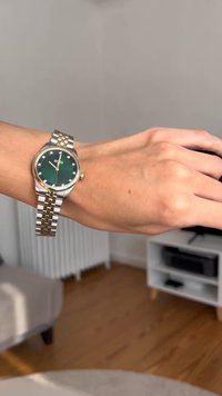 Ümmargune kell, millel on roheline nägu, roostevabast terasest ja kuldse tooniga rihm, hõbedased tunni markerid ja käed.