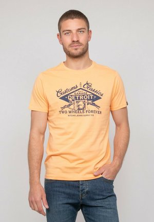 T-shirt en coton orange à manches courtes avec un imprimé graphique bleu comprenant du texte et des motifs de motos. Encolure ras du cou et coupe droite.