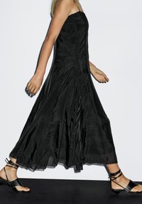 Sort, ærmeløs maxikjole med en flydende silhuet, lavet af glat stof, med subtil tekstur og lagdelt kant. Sammen med sorte, snørede sandaler.