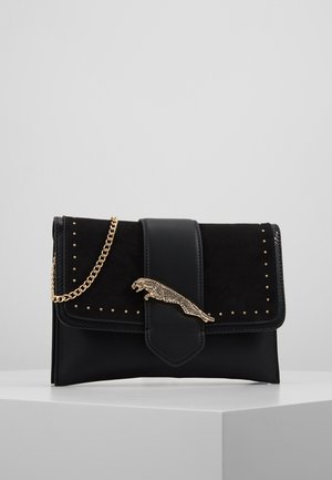 Černé kožené a semišové psaníčko se zlatou sponou ve tvaru jaguára, malými zlatými cvoky a zlatým řetízkovým popruhem na bílém povrchu.