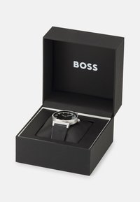 Juodas laikrodis juodame dėkle, su sidabriniu korpusu, juodu ciferblatu ir tekstūruota juoda odine apyranke. "BOSS" logotipas įspaustas dėklo viduje.