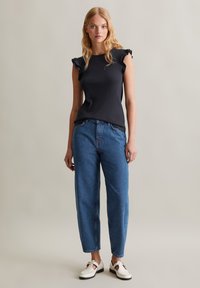 Zwarte geribde mouwloze top met ruches, gecombineerd met lichtblauwe relaxte jeans en witte schoenen met gouden hardware-details.