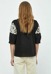 Chemise noire avec des manches bouffantes rehaussées de dentelle, affichant une coupe décontractée et des détails de boutons sur les manches, associée à un pantalon de couleur claire.