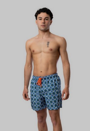 Giovane uomo con capelli corti e ricci che indossa pantaloncini da bagno blu con motivo e coulisse arancione, in piedi davanti a uno sfondo grigio uniforme.