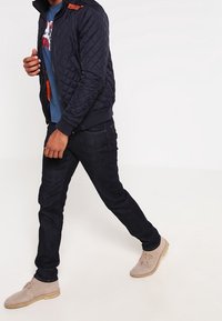 Chaqueta bomber acolchada azul marino con puños de punto, combinada con vaqueros oscuros y zapatos beige con cordones. La chaqueta presenta un detalle de logo en el hombro.