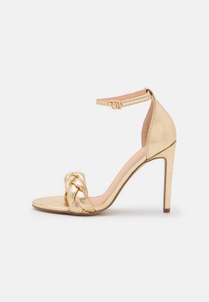 Guld højhælede sandal med flettet frontrem og ankelrem med spænde på ensfarvet hvid baggrund.